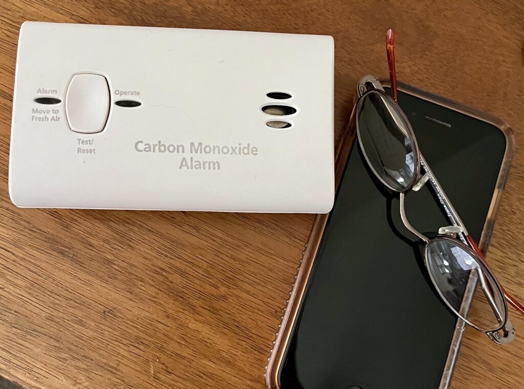 A small travel-size carbon monoxide detector on a table next to a cellphone to show similar size. 